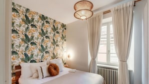 Una cama bien hecha con ropa blanca y una almohada marrón, una pared con estampado floral, una lámpara de fibra natural y una ventana con cortinas.