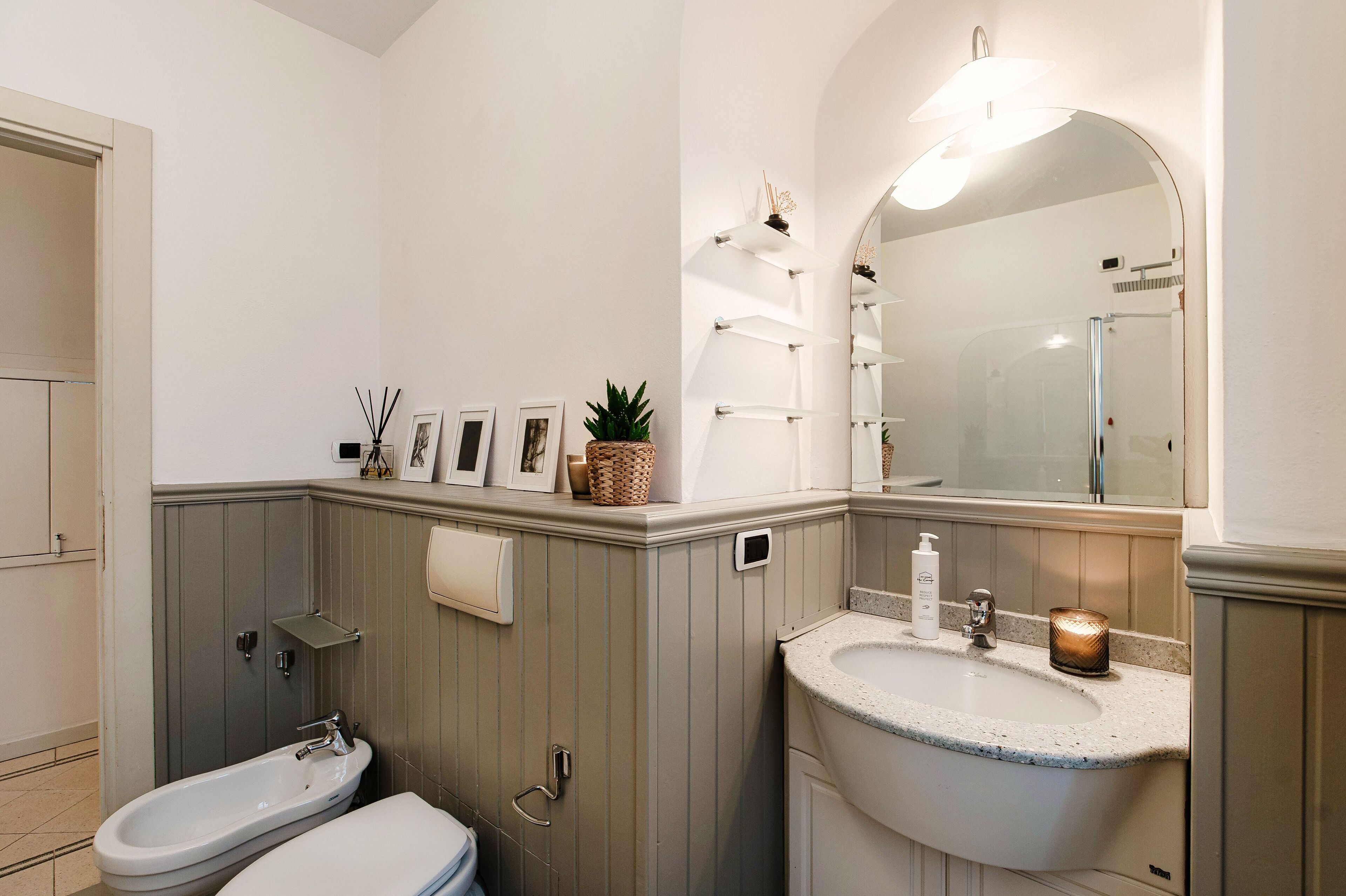 A bathroom with a white sink, a bidet, a mirror, and a shelf with decorative items.
