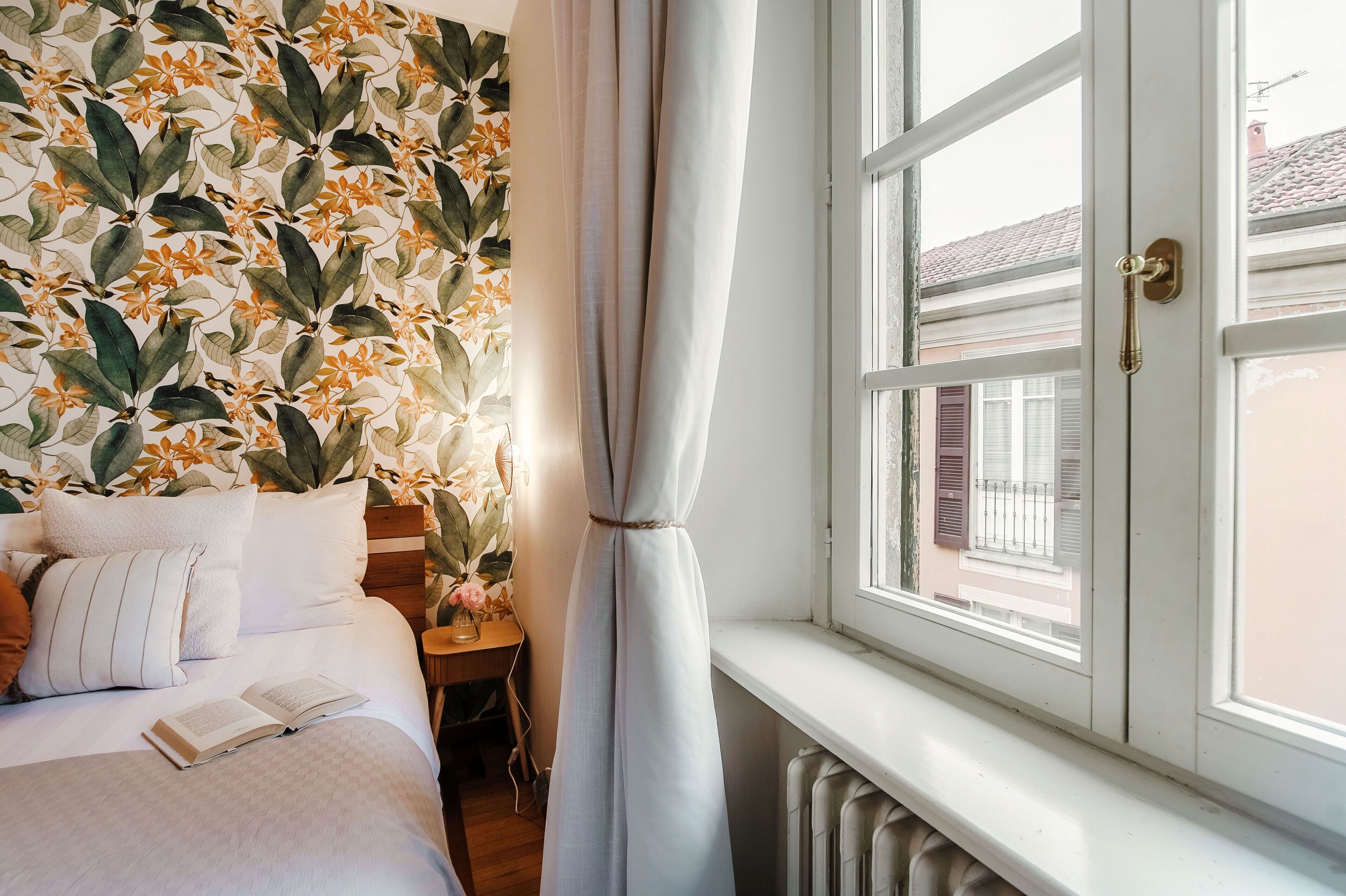 A bedroom with a bed, a nightstand, a window with curtains, and a wall with a leafy pattern.