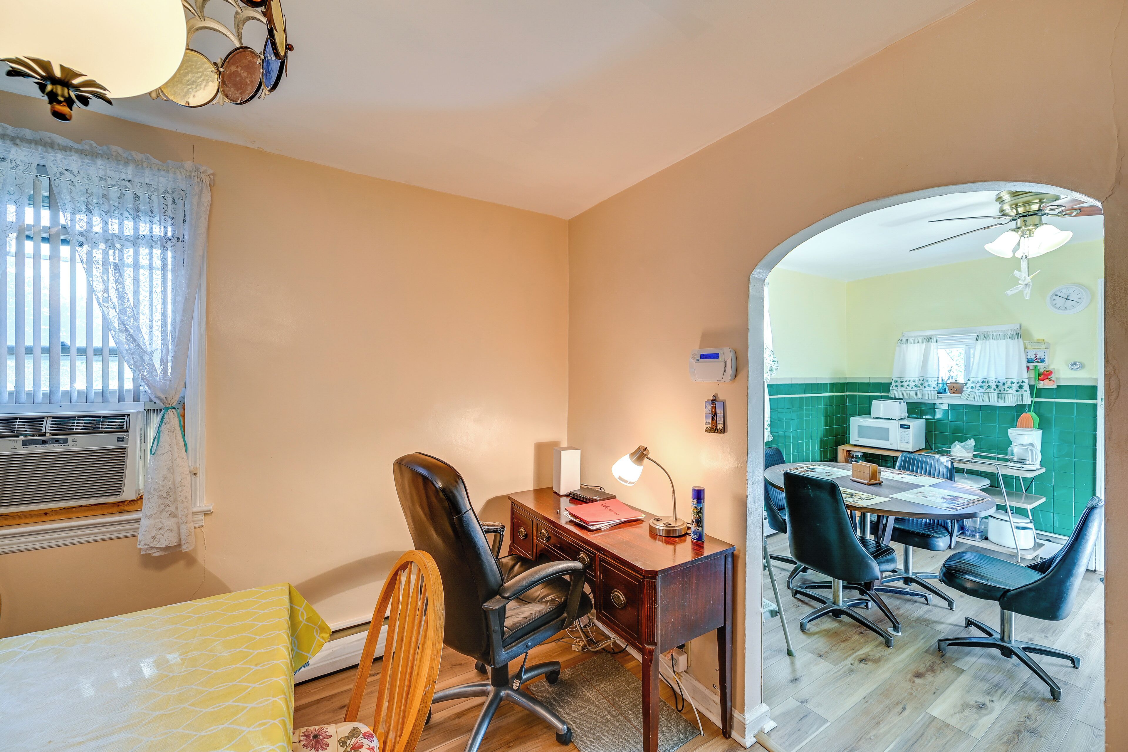 Een kamer met een bureau, stoelen, een bed, een raam met zonwering en een keukenruimte zichtbaar door een boog.