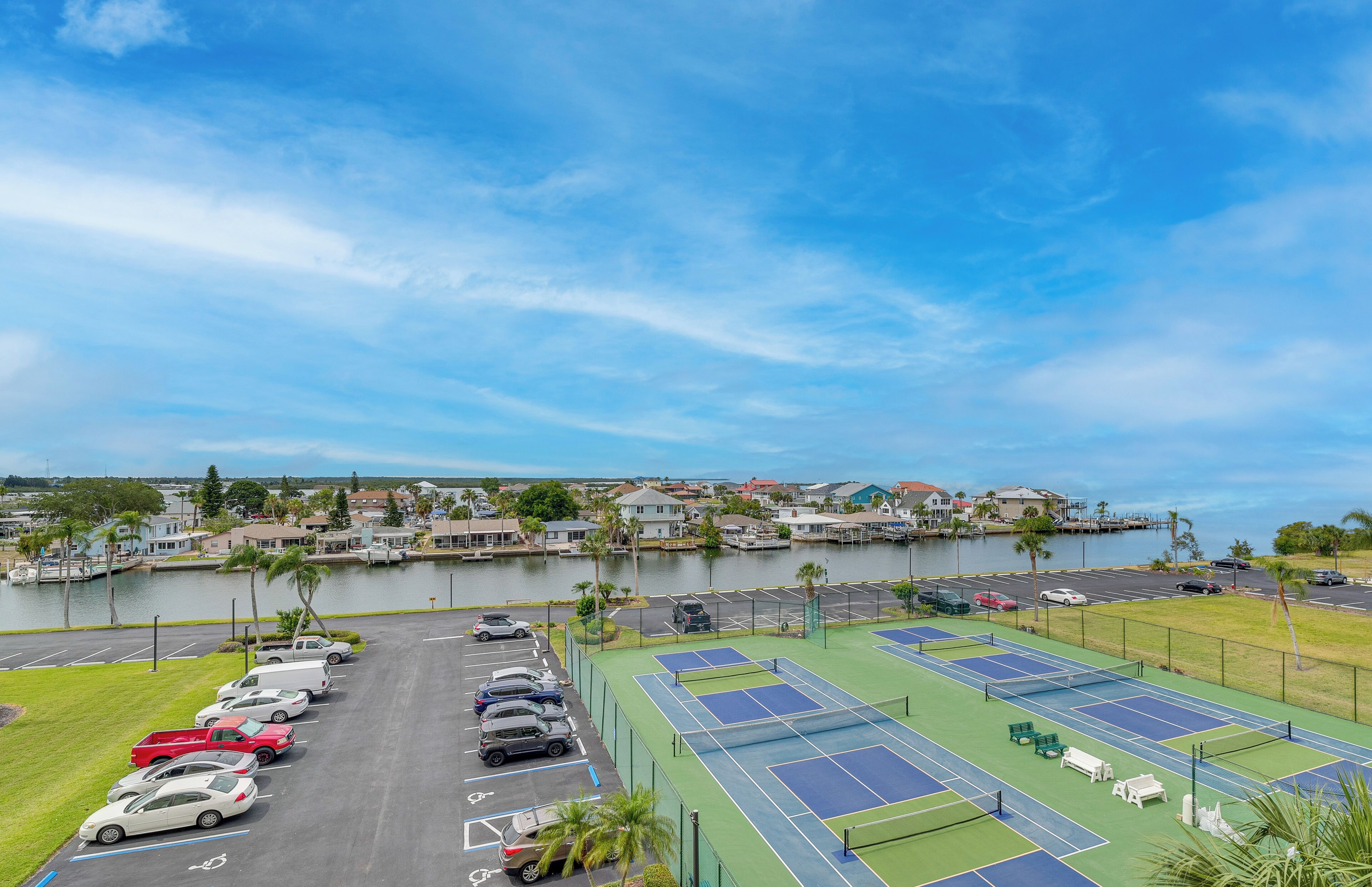 A tennis court with a parking lot in front, a body of water, and houses in the background.