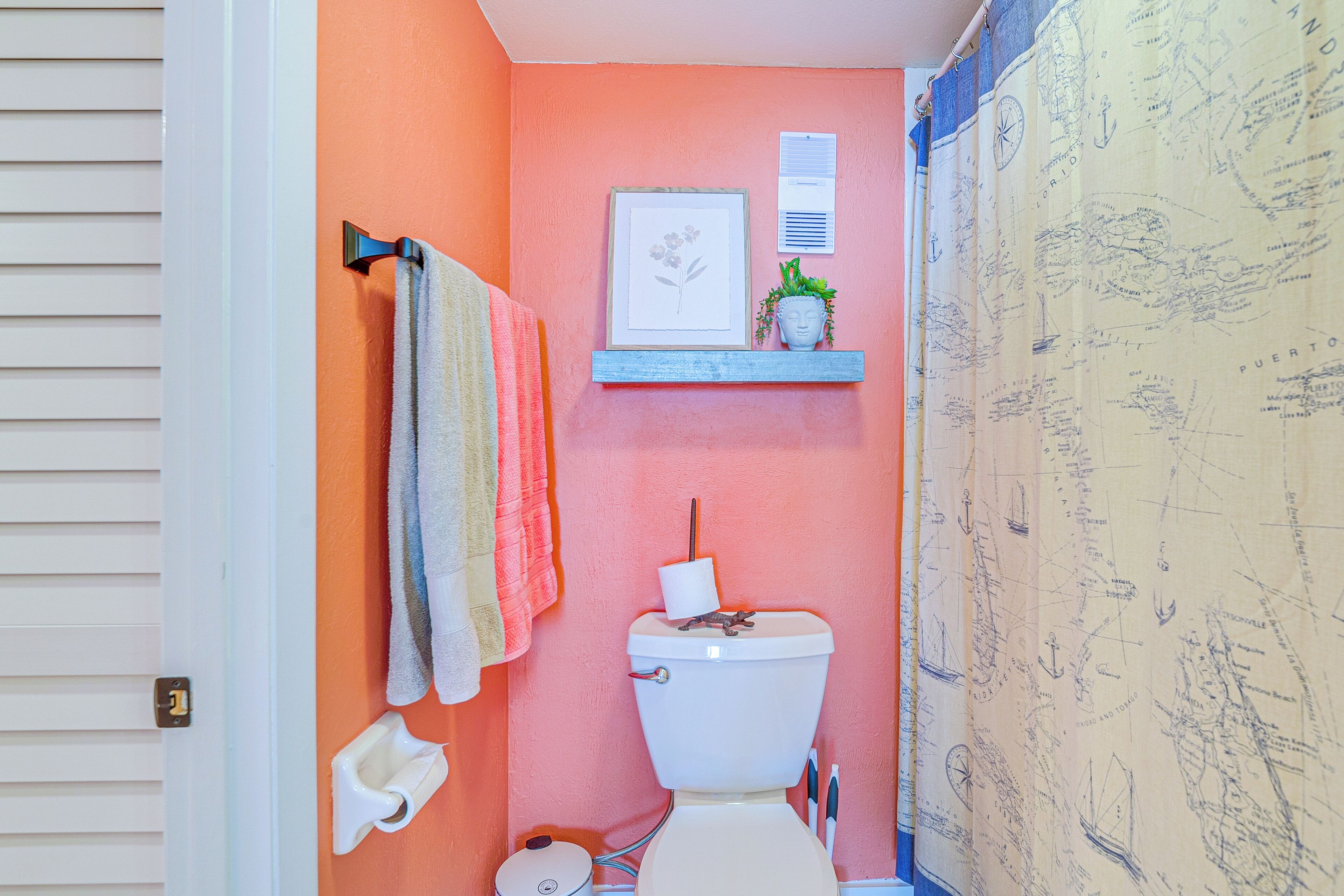 A bathroom with a toilet, a towel rack with multiple towels, a framed picture, a small shelf with a plant, and a decorative curtain with a nautical theme.