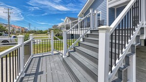 Une terrasse en bois avec une balustrade blanche menant à une maison à l’extérieur grise et dotée d’un escalier blanc.