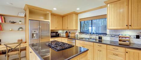 A modern kitchen with wooden cabinets, a stainless steel refrigerator, a built-in stove, and a window with a view of trees.