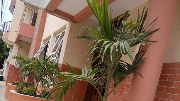 A building with a brick facade, a curved entrance with steps, and potted plants with green foliage and pink flowers. - Résidence Mad 75 (Dakar)