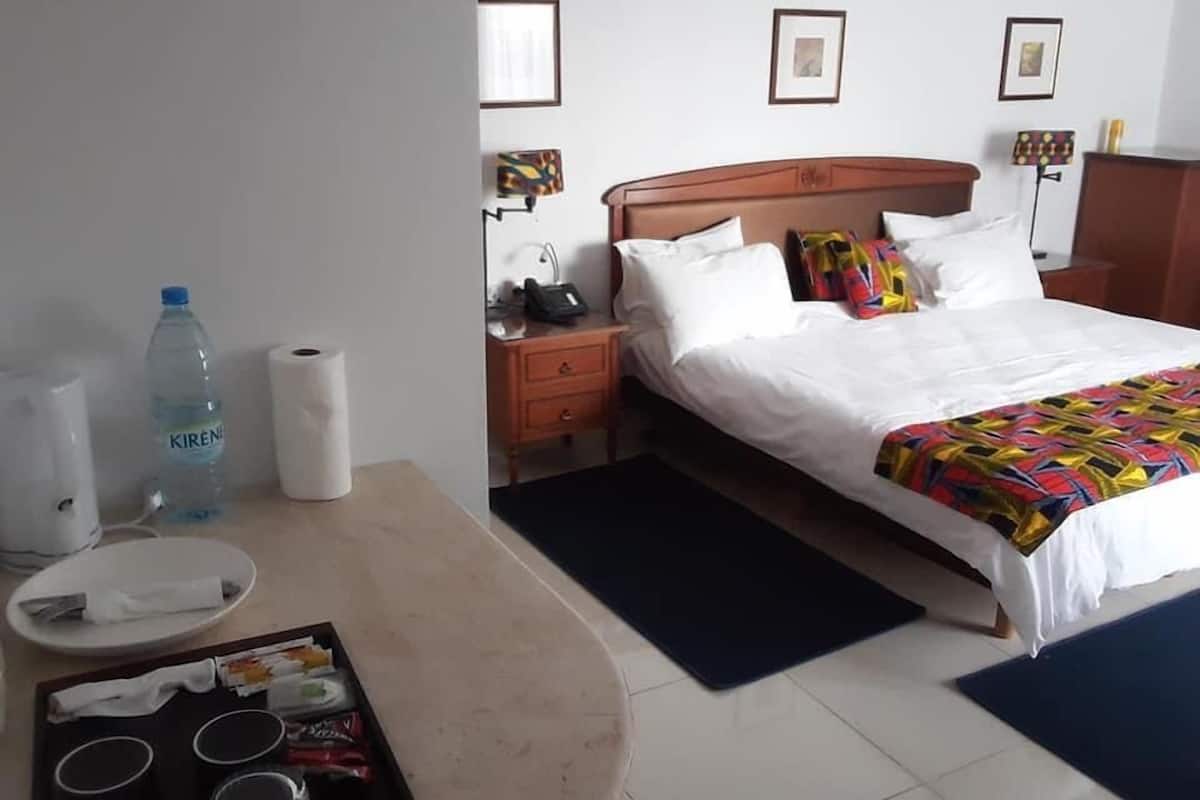 A hotel room with a bed, bedside table, and a desk with a tray of glasses and a water bottle.
