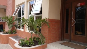 A building entrance with a brown door, two windows with white frames, and a small garden area with plants. - Résidence Mad 75 (Dakar)