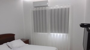 A bedroom with a bed, a nightstand, a TV, and a window with sheer curtains. - Résidence Mad 75 (Dakar)