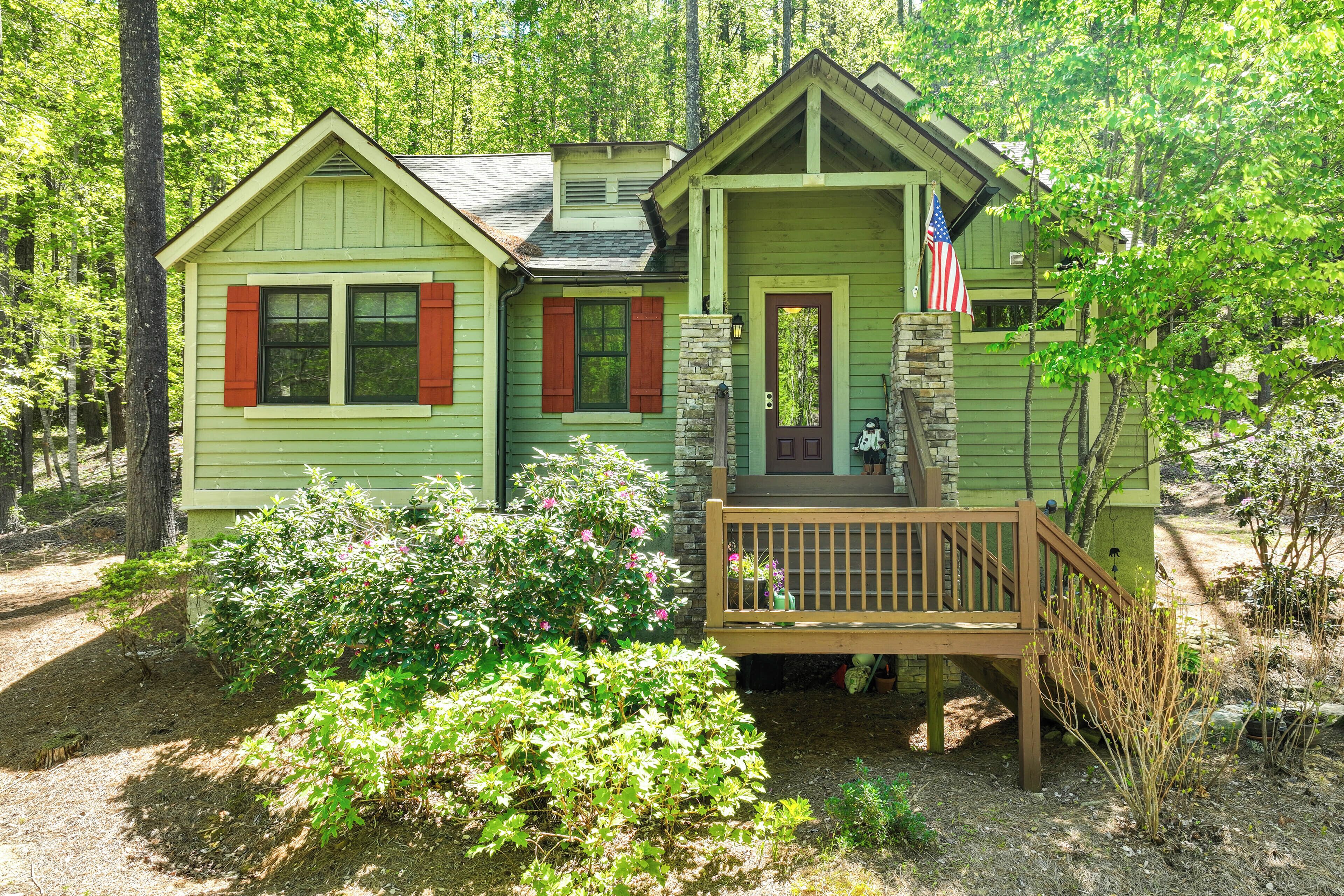 Une petite cabane verte, dotée d’un porche et d’une terrasse en bois, entourée d’arbres et d’arbustes.