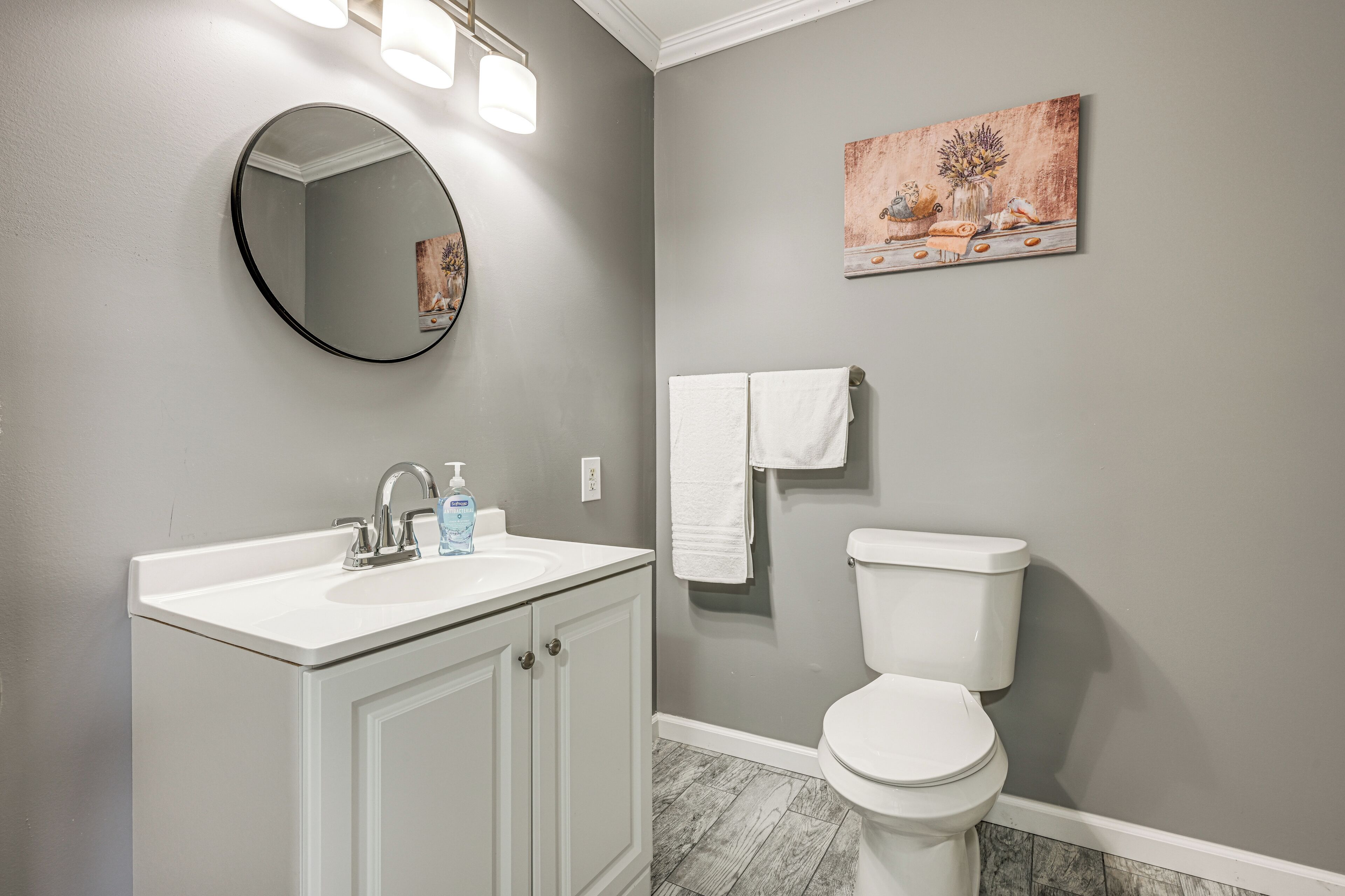 A bathroom with a white vanity, a round mirror, a toilet, and a painting on the wall.