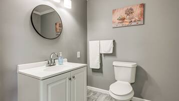 A bathroom with a white vanity, a round mirror, a toilet, and a painting on the wall.
