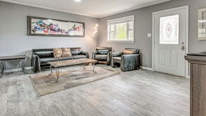 Ein Wohnzimmer mit grauem Sofa, zwei Ledersesseln, einem Couchtisch und einem Teppich. An der Wand hängt ein Gemälde und es gibt ein Fenster mit einer Tür.