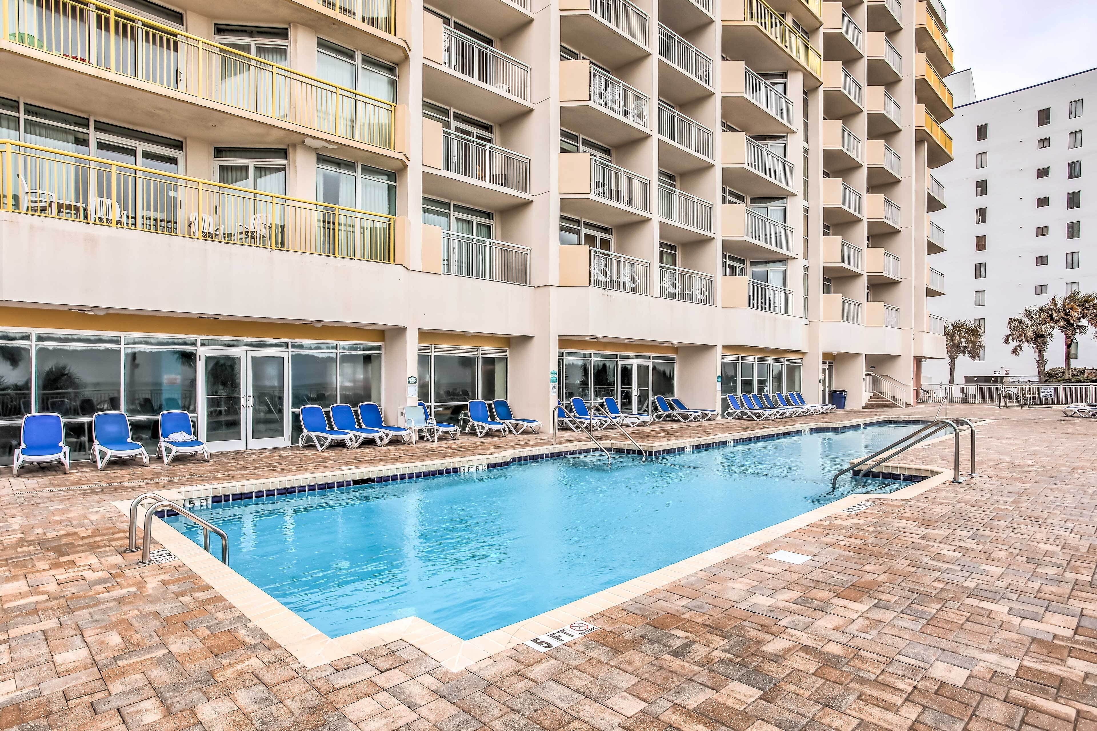 A swimming pool with blue chairs and a brick floor, surrounded by a multi-story building with balconies.
