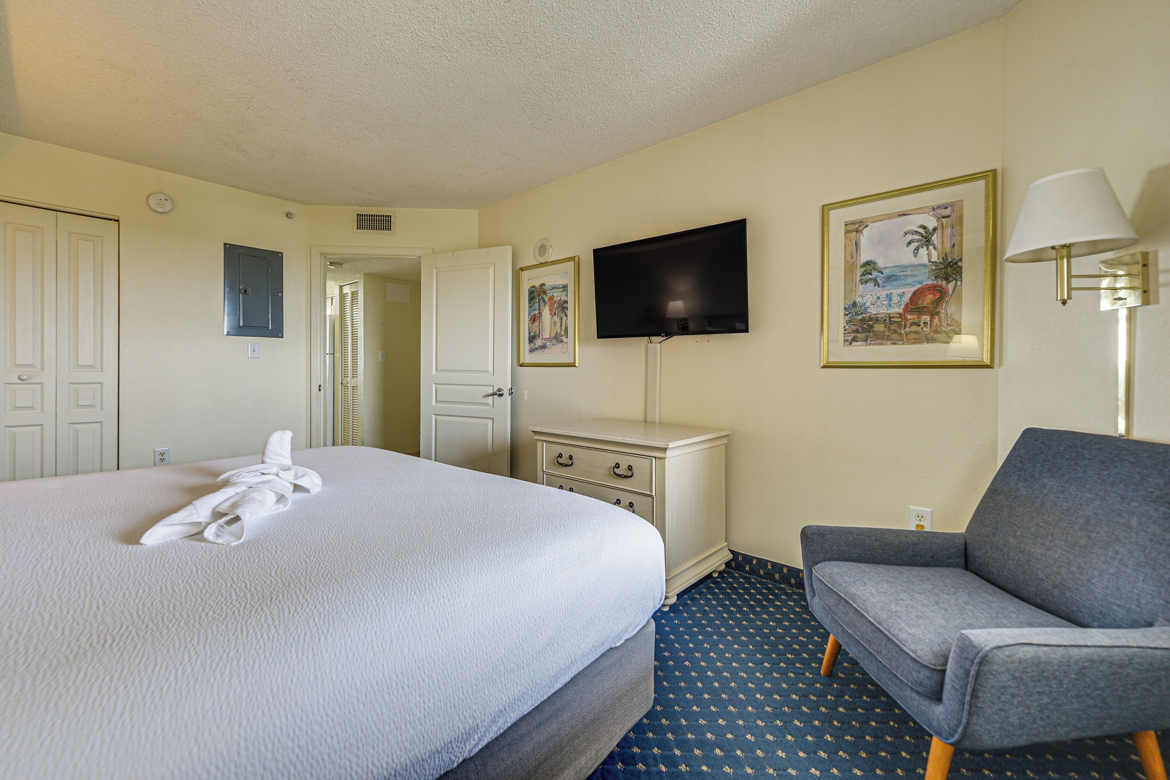 A hotel room with a large bed, a flat-screen TV mounted on the wall, a desk with drawers, a grey armchair, and a patterned carpet.