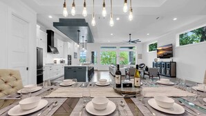 Een moderne keuken met witte kasten, een eethoek voor zes personen met een glazen tafel, en een groot raam met uitzicht op groen.
