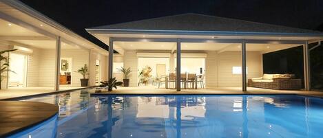 A modern house with a swimming pool, large glass doors leading to a patio, and a well-lit interior.