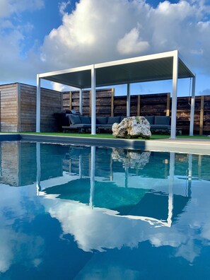 A modern outdoor pool area with a wooden deck, a covered seating area, and a large rock centerpiece. - Nakolodge 4 chambres - piscine / balneo / jardin privatifs  (Sainte-Anne)