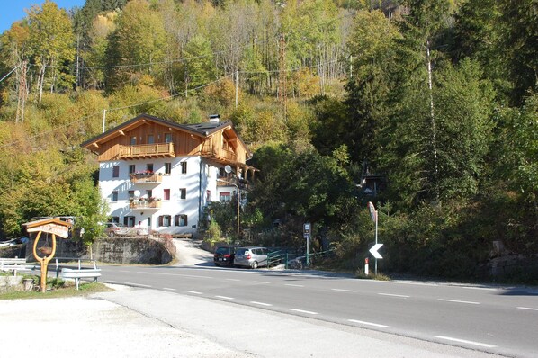A two-story wooden building with balconies, a road in front, and a forested hillside. - COZY AND SPACIOUS ALPINE ATTIC ROOM WITH PETS ALLOWED (Alleghe)
