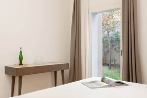 A room with a bed, a table with a bottle and a glass, and a view of a garden through a window.
