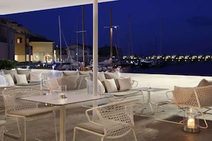 An outdoor dining area with wicker furniture and tables, overlooking a marina with boats and buildings in the background.