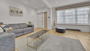A modern living room with a grey sofa, a glass coffee table, a large window with blinds, and a radiator. - Family Town-House Behind Park Lane (London)