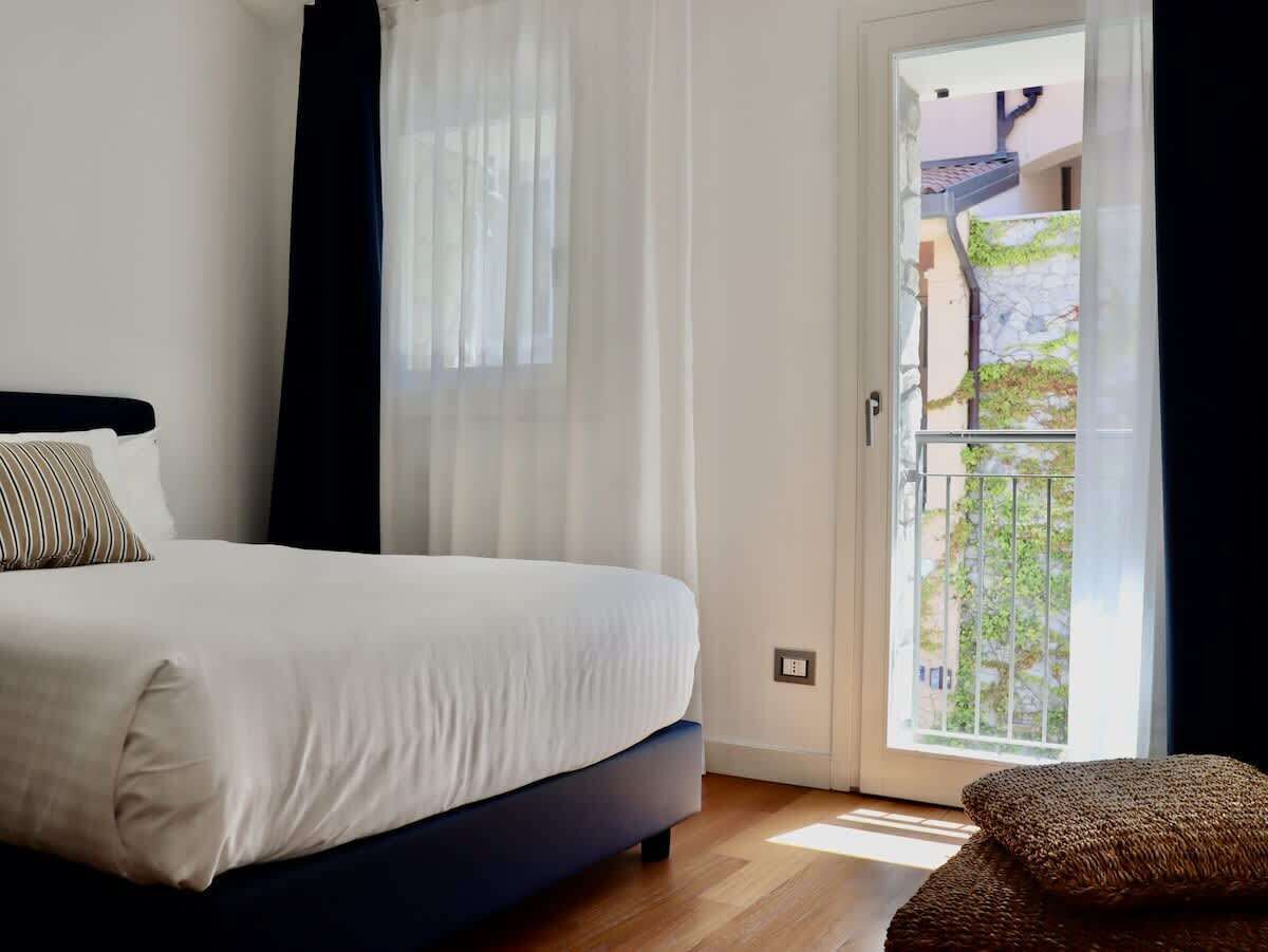 A bedroom with a bed, a window with curtains, a balcony door, and a view of greenery outside.