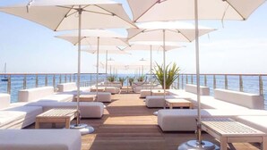 Eine Terrasse mit weißer Gartenmöbelgruppe, Sonnenschirmen und Blick aufs Meer.