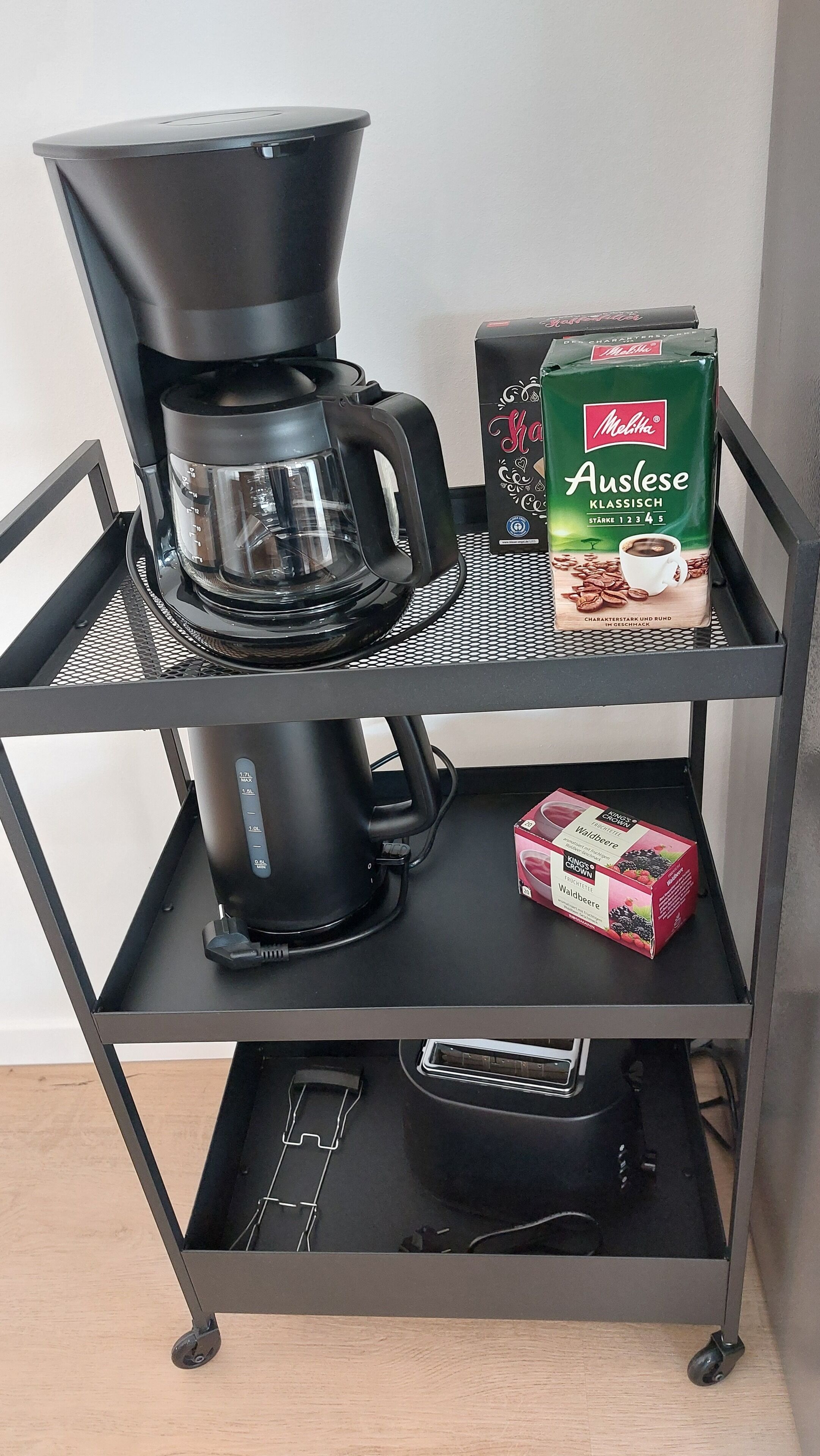 Uma cafeteira em cima de um carrinho de metal, com duas caixas de café e um sachê de café.