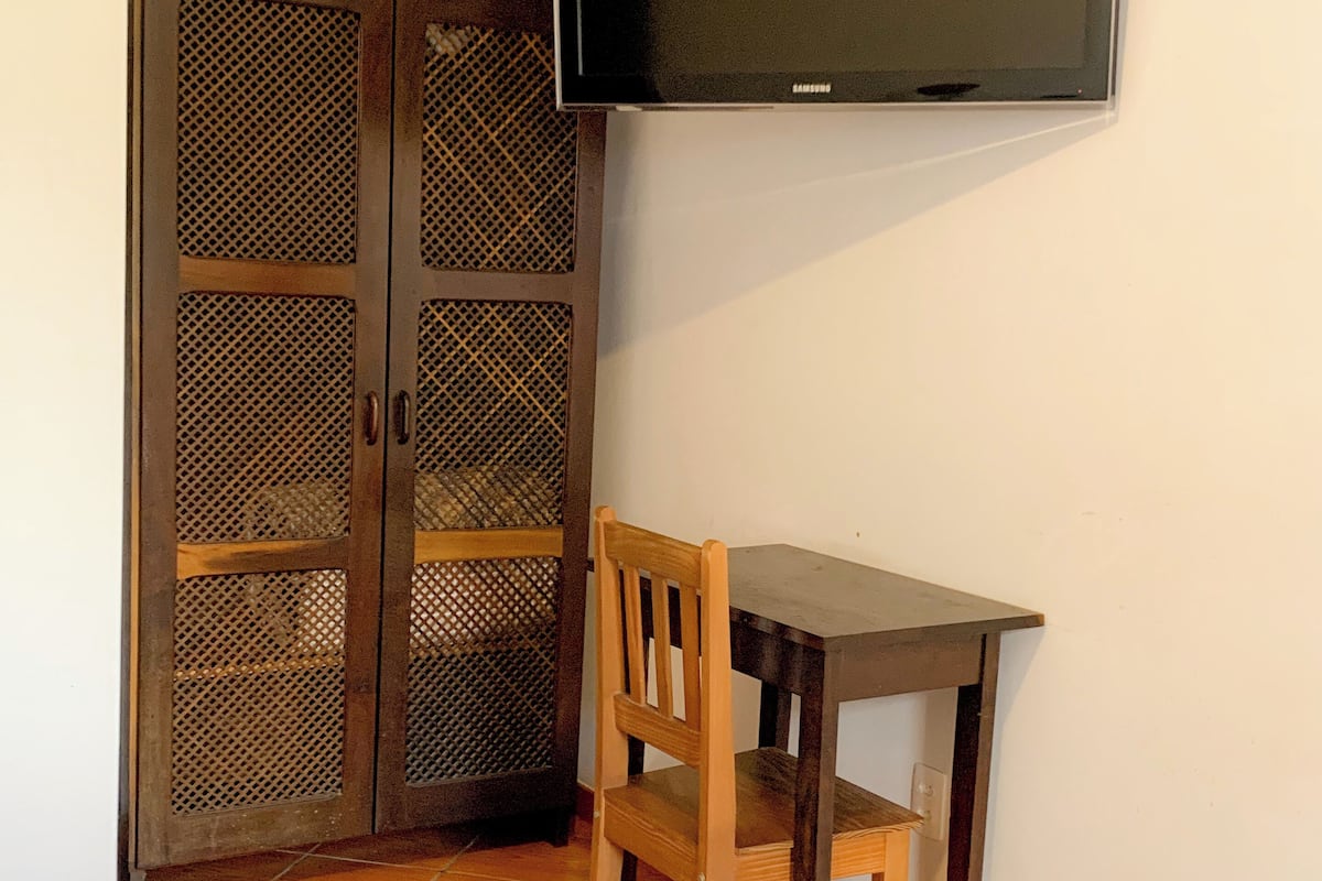 Ein Schlafzimmer mit einem Bett, einem Holztisch, einem Stuhl, einem an der Wand montierten Fernseher und einem Kleiderschrank mit Drahtgittertür.