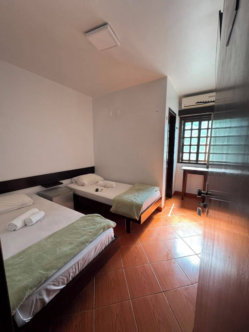 A hotel room with two beds, a desk, a window with blinds, and a built-in air conditioning unit.