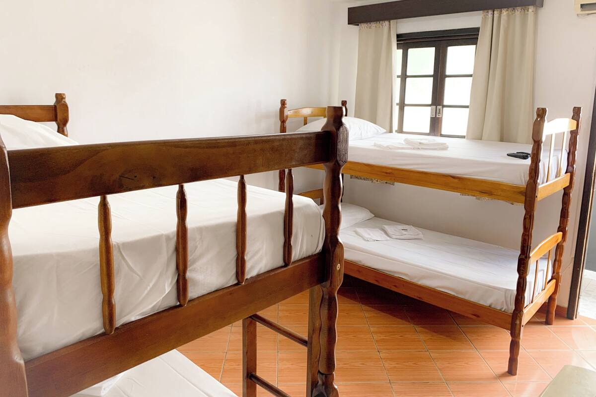 Ein Schlafsaalzimmer mit drei Etagenbetten aus Holz, weißer Bettwäsche, einem Fenster mit Vorhängen und einem gefliesten Boden.