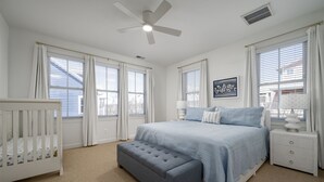 Ein Schlafzimmer mit einem Bett, einem Nachttisch, einem Babybett, einem Deckenventilator und großen Fenstern mit Jalousien.