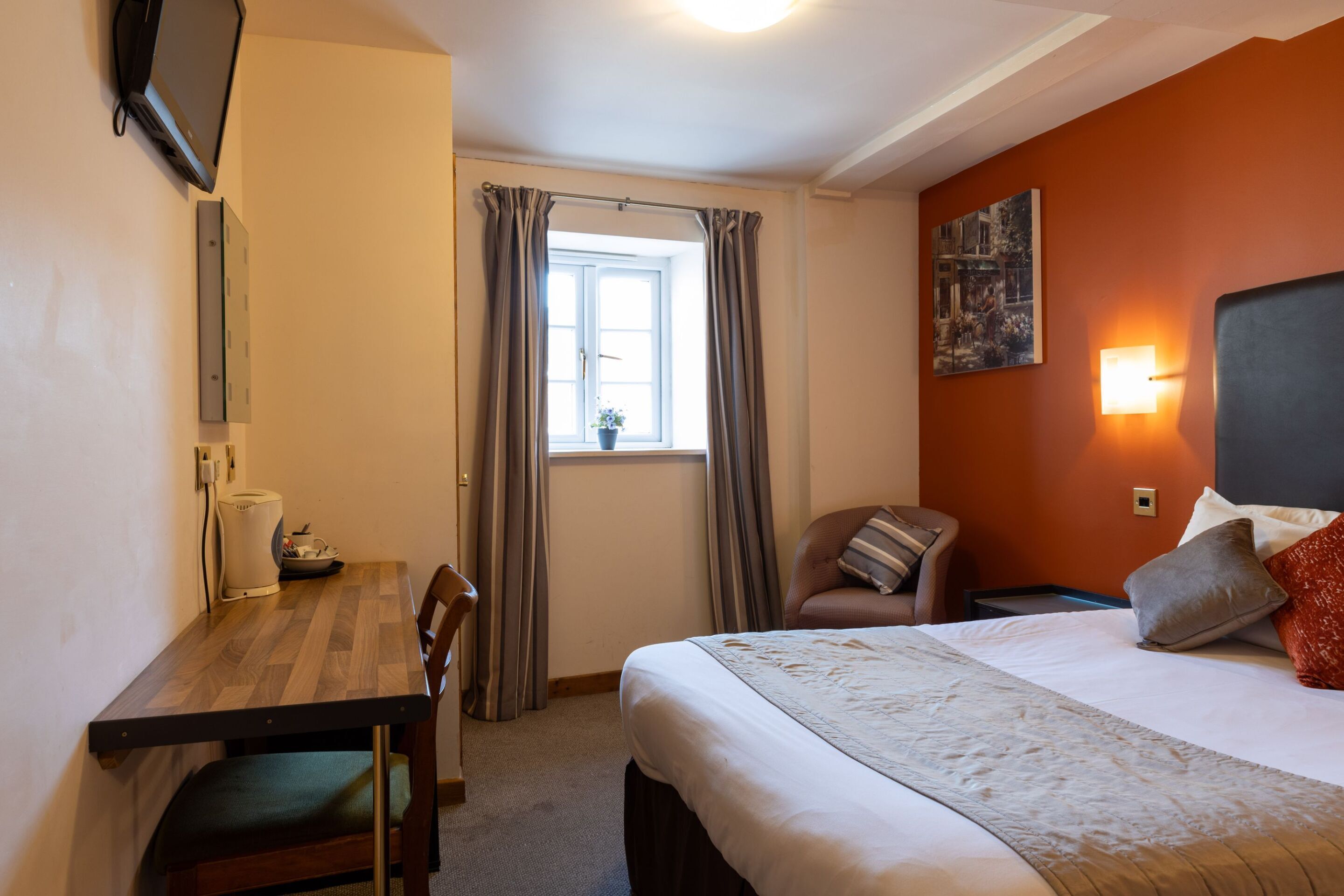 A hotel room with a bed, a desk, a chair, a window with curtains, a wall-mounted TV, and a painting.