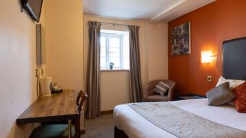 A hotel room with a bed, a desk, a chair, a window with curtains, a wall-mounted TV, and a painting.