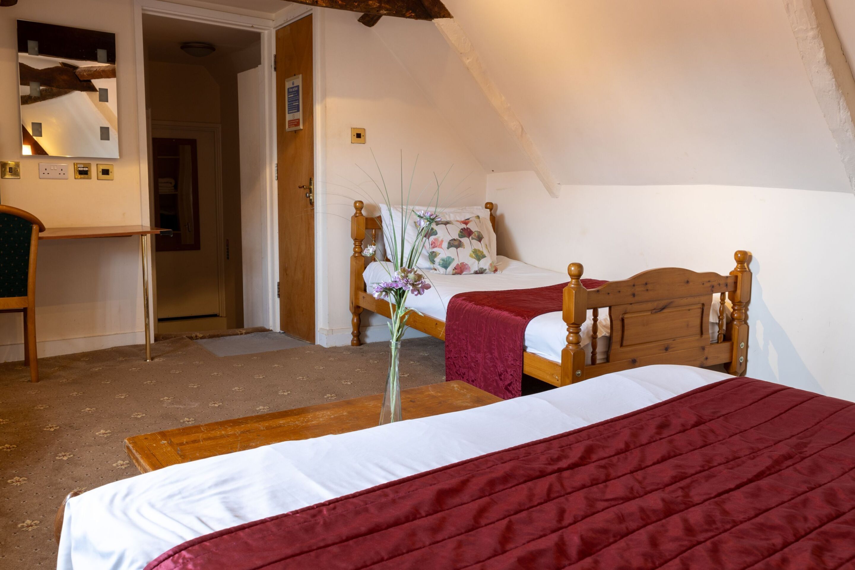 A bedroom with a wooden bed, a red bedspread, a wooden nightstand, and a mirror on the wall.