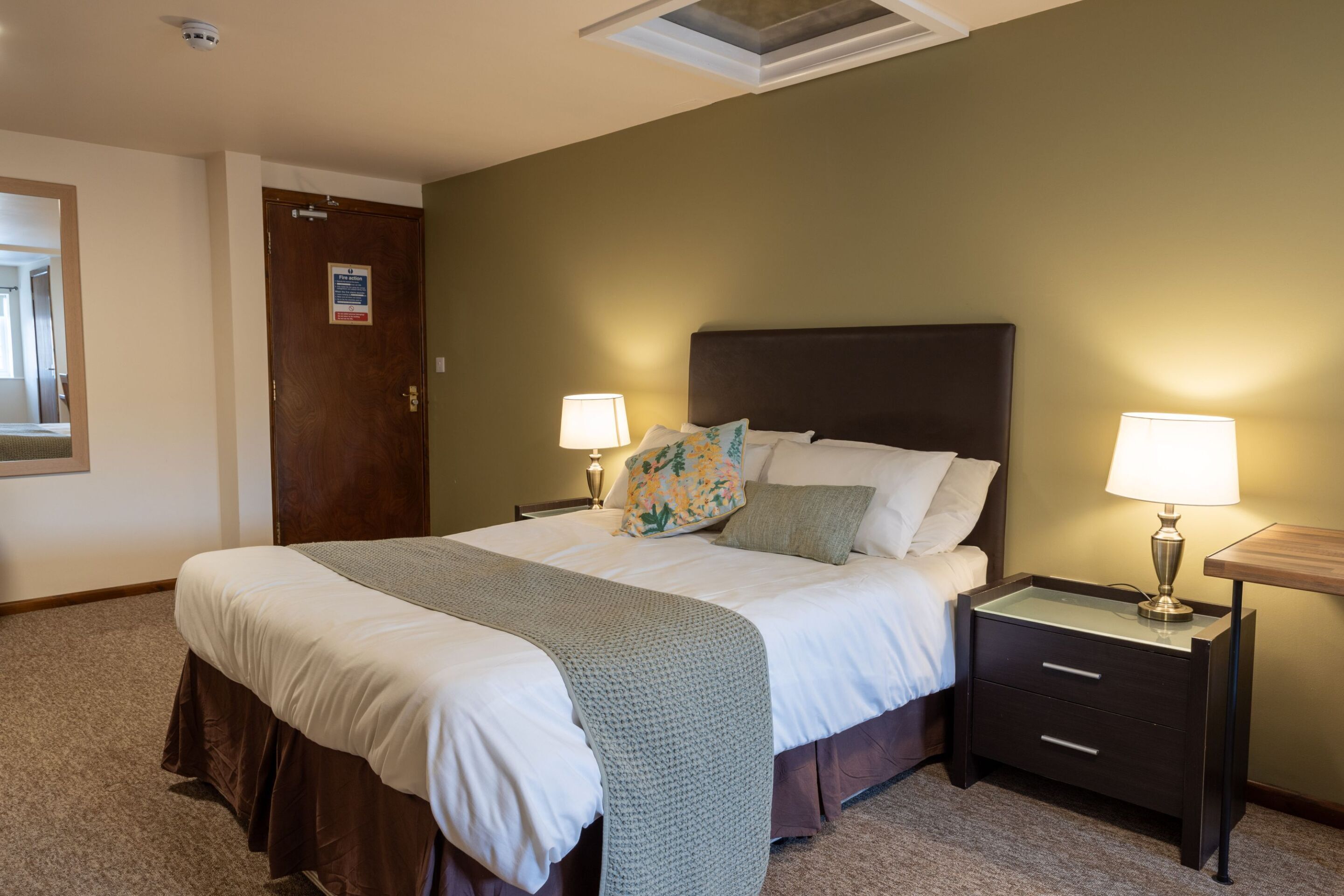 A neatly made bed with a grey blanket, two bedside tables with lamps, and a wooden door with a sign.