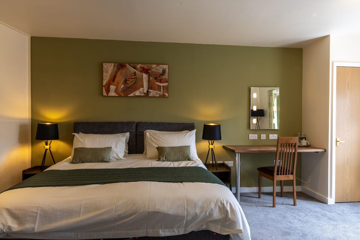 A hotel room with a large bed, two bedside lamps, a desk with a chair, a mirror, and a painting on the wall.