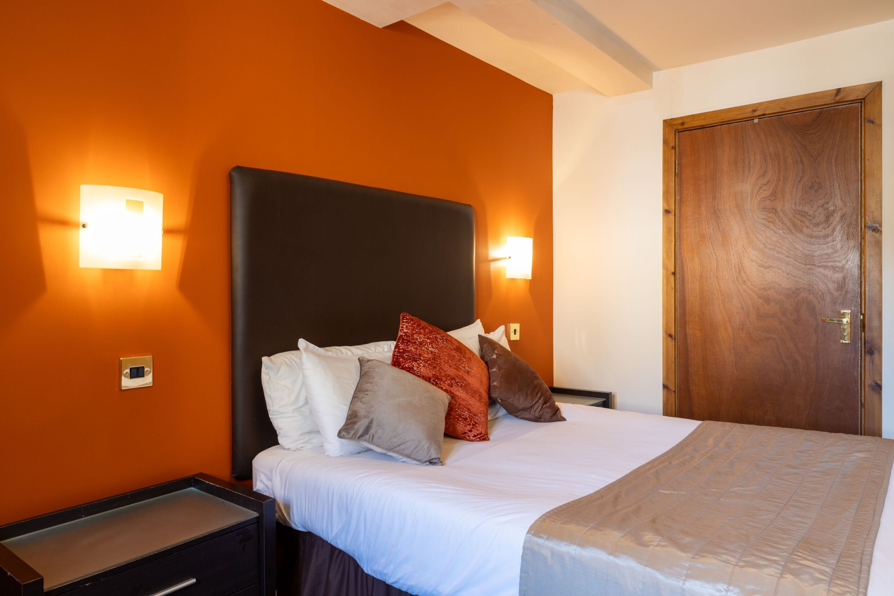 Una camera d'albergo con un letto, due comodini, una porta in legno e luci a parete.
