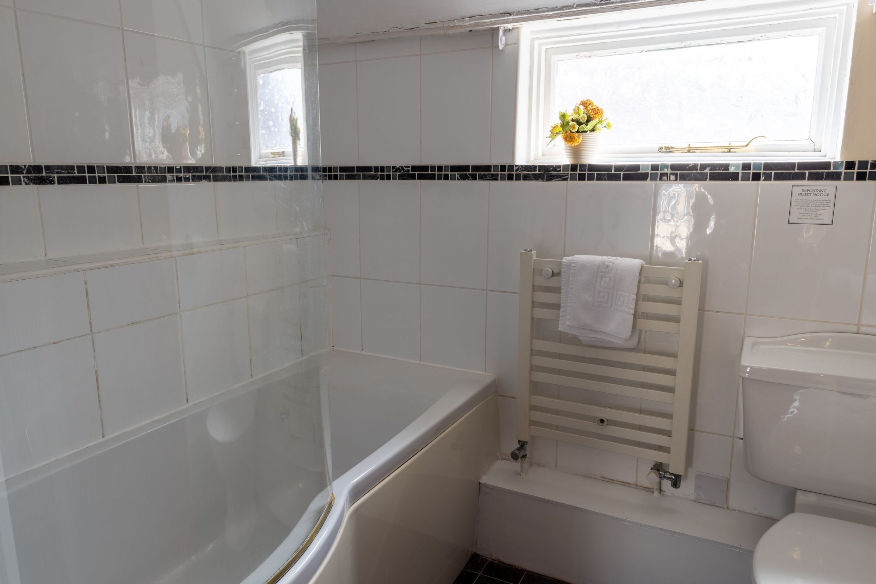 A bathroom with white tiles, a bathtub, a toilet, and a window with flowers.