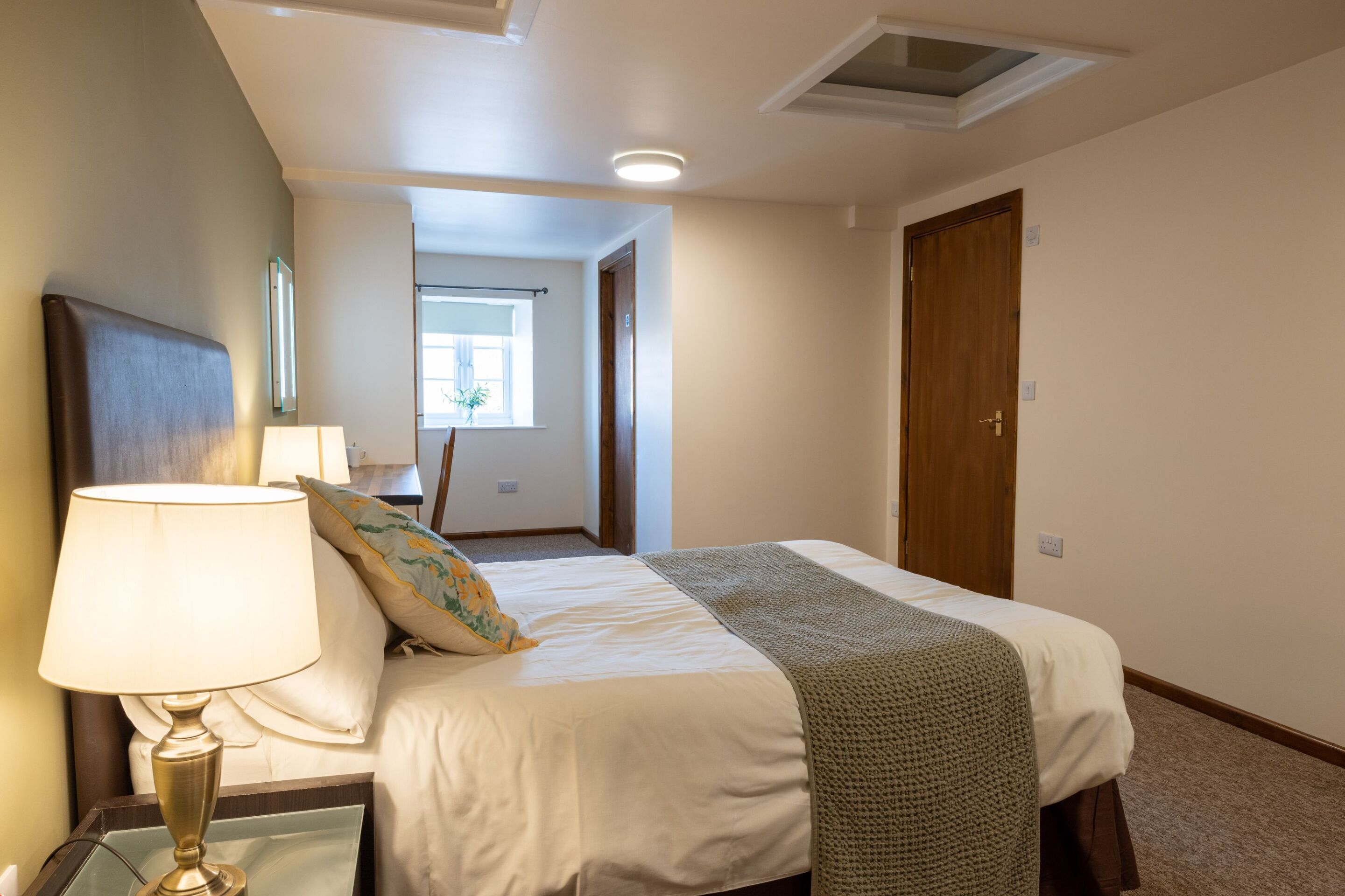 A hotel room with a bed, a nightstand with a lamp, a window, and a door.