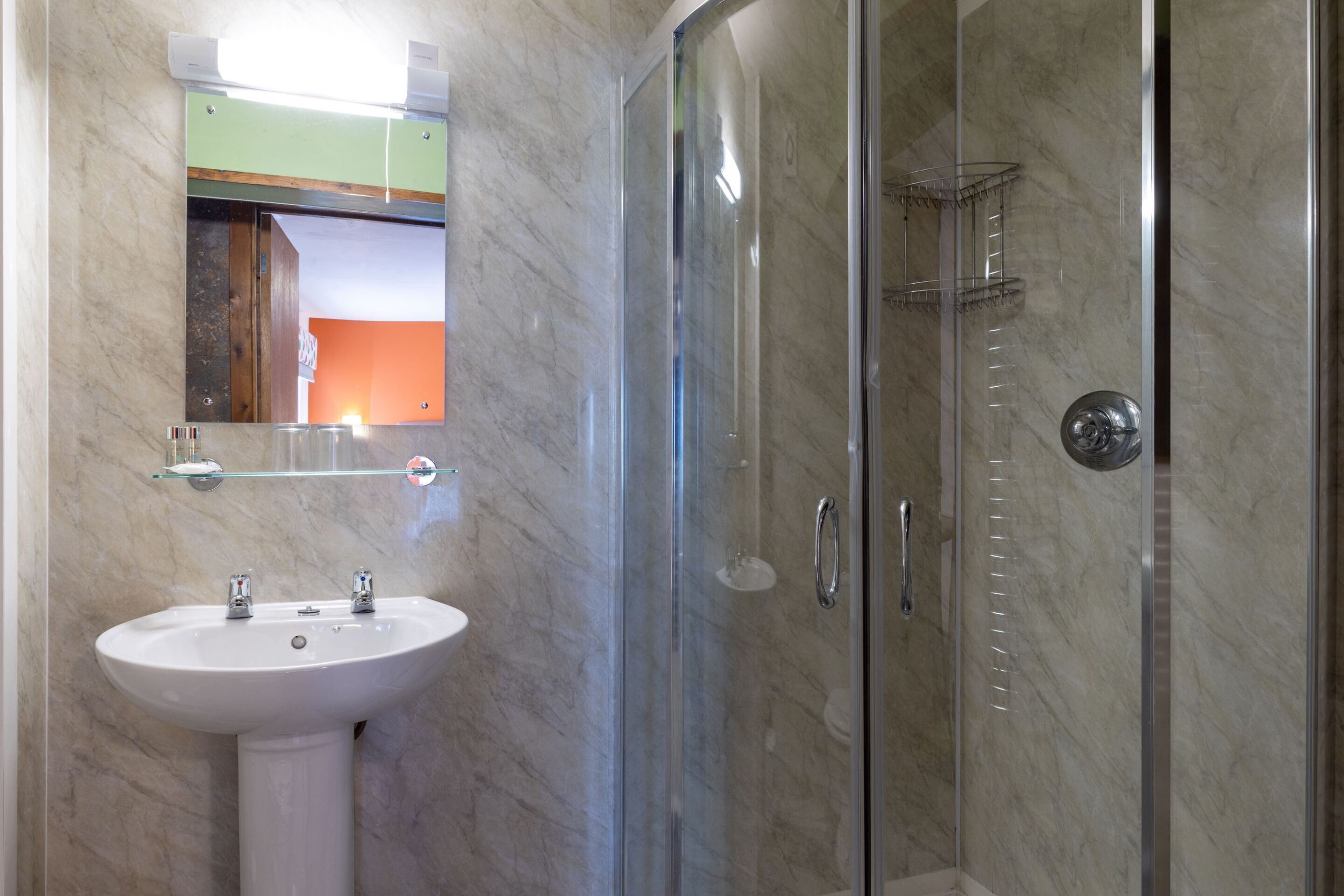 Une salle de bain avec un lavabo sur colonne blanc, une cabine de douche en verre et un miroir au-dessus du lavabo.