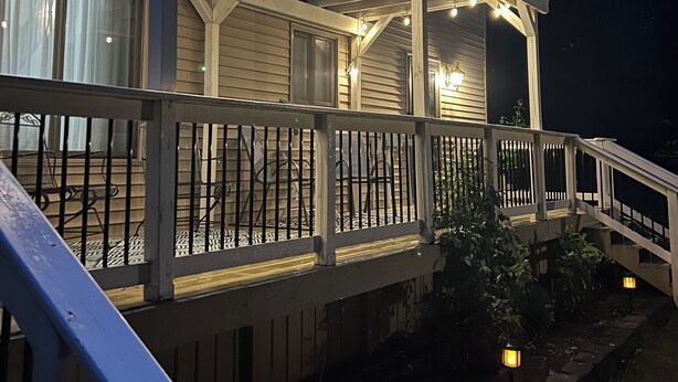 A covered outdoor deck with string lights, a wooden railing, and a stone pathway leading to a house. - Secord Lake Private Wooded Luxury Retreat (Alger)