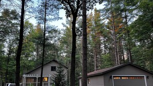 A cabin with a porch and a garage, surrounded by tall trees. - Secord Lake Private Wooded Luxury Retreat (Alger)