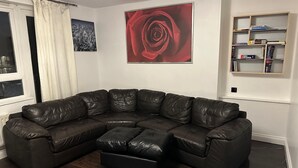 Un salon comprenant un canapé d’angle en cuir marron foncé, un pouf noir, un tableau représentant une rose rouge, une estampe représentant un paysage urbain et une bibliothèque remplie de livres et ornée d’un globe terrestre.