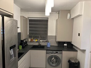 A compact kitchen with a stainless steel refrigerator, a built-in washing machine, and a sink with a window above it. - Stylish 1BR Apt w/ Fireplace & High-speed Wifi (London)