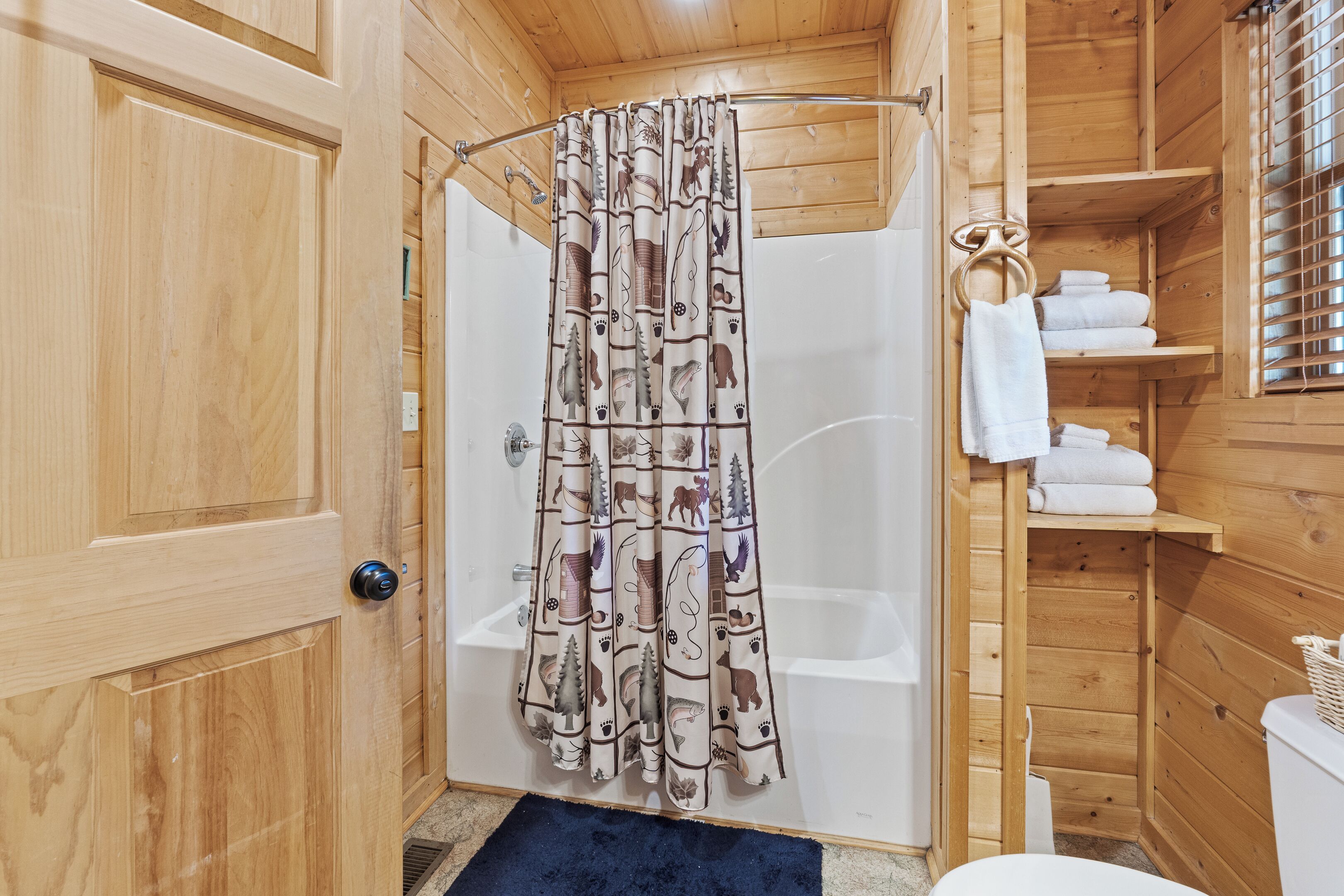A wooden cabin bathroom with a shower curtain featuring animal prints, a white bathtub, and a wooden shelf with towels.