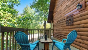 Um deck de madeira com duas cadeiras Adirondack azuis e uma mesa de madeira, cercado por um muro de madeira e árvores.
