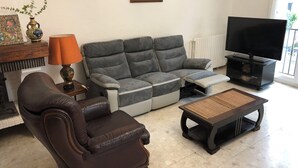 A living room with a sofa, a brown leather armchair, a television on a stand, and a small table with a woven top. - Grande Maison de Village à Estagel (ESTAGEL)