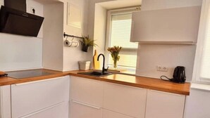 Een kleine keuken met witte kastjes, een houten aanrecht, een zwarte kraan en een raam met zonwering.