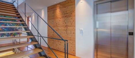 A modern staircase with wooden steps and metal railings, leading to a room with a wooden panel wall and a metallic elevator.